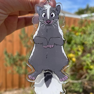 May include: A clear acrylic keyring with a cartoon skunk design. The skunk is depicted in grey and white, with pink details on its paws and nose. It has a silver metal ring for attaching to keys or bags.