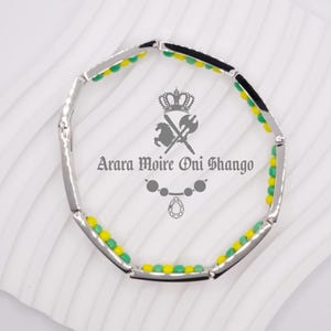 May include: A silver bracelet with green and yellow beads. The bracelet has a black enamel band with a crown and crossed swords design. The text "Arara Moire Oni Shango" is printed on the band.