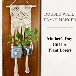 Può includere: Un appendiabiti da parete doppio in macramè con due vasi, uno azzurro chiaro e uno grigio scuro, ciascuno con una pianta verde. Il macramè è color crema e pende da un tassello di legno. Il testo recita "DOUBLE WALL PLANT HANGER" e "Mother's Day Gift for Plant Lovers."