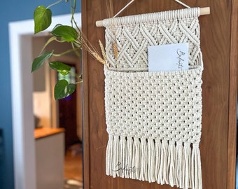 Op de afbeelding: Crèmekleurige macramé wandhanger met een geometrisch patroon en een zak. De hanger heeft franjes aan de onderkant en een houten deuvel aan de bovenkant. Een kaart met het woord "Belarts" zit in de zak. Tarwearen zijn bevestigd.