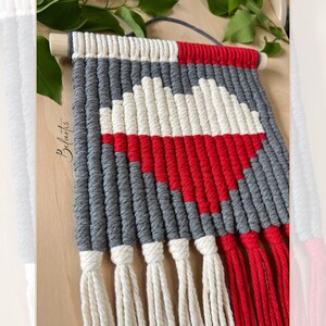 Op de afbeelding: Een grijze, witte en rode macramé wandhanger met franjes. De wandhanger is gemaakt van katoenen koord en heeft de vorm van een hart.