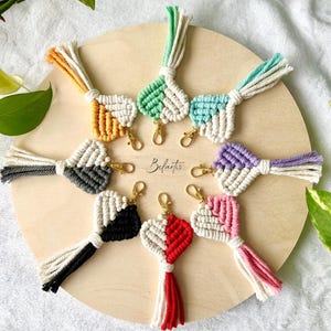 May include: A collection of handmade macrame heart-shaped keychains in various colors, including orange, green, blue, purple, pink, red, black, and gray. Each keychain features a gold-colored clasp and a tassel. The keychains are arranged on a wooden circle.