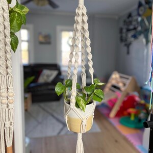 Puede incluir: Un macetero de macramé con un anillo de madera en la parte superior y una pequeña planta en maceta colgando de él. El macetero está hecho de cordón de algodón de color natural y tiene cuentas de madera.