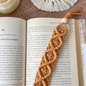 May include: A close-up of a mustard yellow macrame bookmark with a wooden bead at the top. The bookmark is resting on an open book with text about mental subtraction. The text on the book reads "Mental Subtraction: Benefits".
