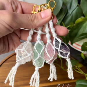 Puede incluir: Cuatro dijes de llavero de cristal con cierres dorados. Cada dije está envuelto en macramé blanco y tiene una borla blanca. Los cristales son transparentes, verdes, rosas y morados.