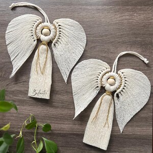 May include: Two white macrame angel ornaments with gold accents and wooden beads. The angels have long, flowing wings and a tassel-like body. The ornaments are hanging from a wooden surface.