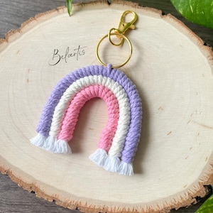 May include: A handmade macrame rainbow keychain in shades of purple, pink, and white, with a gold-colored clasp and ring. The keychain is on a wooden surface, and the word "Belartis" is written in cursive.