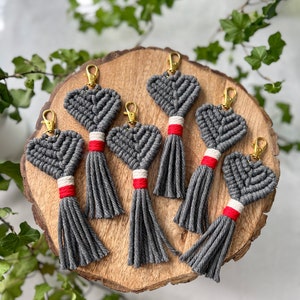 Puede incluir: Seis llaveros de corazón gris con borlas rojas y blancas. Los llaveros están hechos de cuerda y están dispuestos sobre una superficie de madera.