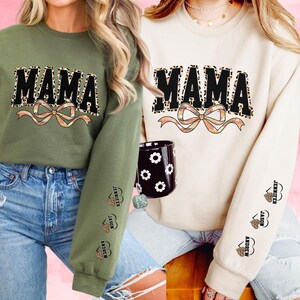May include: Two sweatshirts, one olive green and one cream, each featuring the word "MAMA" in black block letters with a leopard print fill. A bow design is below the word. The sleeves have names printed with heart designs.