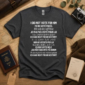 May include: Dark gray t-shirt with the text "I DID NOT VOTE FOR HIM" in white, along with translations in multiple languages. The shirt is displayed on a world map, with a passport and camera nearby.