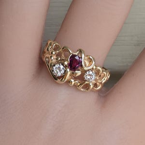 Puede incluir: Un anillo de oro con una piedra preciosa roja oscura de corte cuadrado en el centro, flanqueada por dos piedras preciosas redondas y transparentes más pequeñas. El anillo tiene un diseño calado ornamentado. El anillo está en el dedo de una persona.