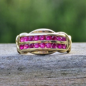 May include: A gold band ring featuring two parallel rows of small, round, vibrant pinkish-red rubies set within a channel. The metal has a polished finish, and the design includes smooth, curved gold segments that frame the gemstone rows.