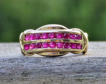 Puede incluir: Un anillo de banda de oro con dos filas paralelas de pequeños rubíes redondos de color rojo rosado vibrante engastados en un canal. El metal tiene un acabado pulido y el diseño incluye segmentos de oro lisos y curvos que enmarcan las filas.