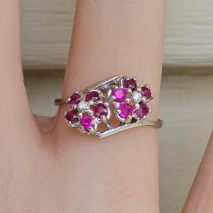 May include: A silver ring with two floral arrangements. Each arrangement features bright pink gemstones and small white gemstones. The ring is displayed on a finger, highlighting its intricate design and sparkling details.