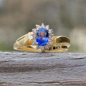 Puede incluir: Un anillo de oro con una gran piedra preciosa azul ovalada, rodeada de piedras más pequeñas y claras. La banda del anillo tiene un diseño dividido. El anillo está sobre un fondo natural borroso.