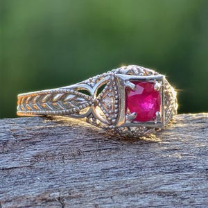 May include: A silver-toned ring featuring an oval-cut vibrant red gemstone set in a square frame. The band displays an intricate, textured leaf-like pattern along the sides, with small, delicate metal beadwork detailing surrounding the central stone.