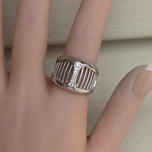 May include: A silver-toned ring with a rectangular design featuring vertical bars and three small, clear gemstones. The ring is worn on a finger, showcasing its intricate details and modern style. The ring's design is reminiscent of a car grill.