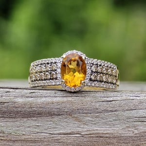 May include: A ring featuring a central oval-cut yellow citrine gemstone surrounded by a halo of small white stones. The band is wide and decorated with multiple rows of small brown stones flanked by additional rows of tiny white stones.