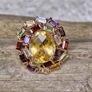 Könnte beinhalten: Ein Ring aus Roségold mit einem großen, oval geschliffenen gelben Citrin in der Mitte, umgeben von einem Kranz aus mehrfarbigen Edelsteinen im Baguetteschliff, darunter violette, grüne, rote und klare Steine in kreisförmiger Anordnung.