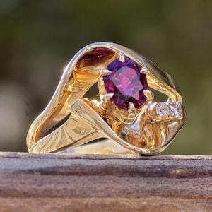 May include: A gold ring with a large, round, dark purple gemstone surrounded by smaller, clear gemstones. The ring has a unique, abstract design.