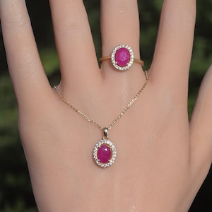 Könnte beinhalten: Ein Goldring und eine passende Goldanhängerkette, jeweils mit einem ovalen, leuchtend rosa Edelstein, der von einem Kranz aus kleinen, klaren weißen Steinen umgeben ist. Das Schmuckset zeigt ein einheitliches Design mit polierten Goldbändern.