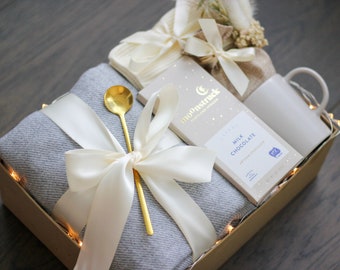 May include: A gift box filled with a gray throw blanket, a gold spoon, a white mug, a bar of milk chocolate, and a small bag of dried flowers. The box is tied with a white satin ribbon and has a string of white lights around the edge.