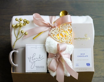 May include: A gift basket filled with items for relaxation and self-care. The basket includes a mug, a white blanket, a pair of socks, a box of artisan chocolate, a box of crackers, a heart-shaped light, and a gold spoon.