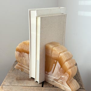May include: A pair of amber-colored onyx bookends with a layered, fan-like design. Two hardback books with off-white covers are held between the bookends. The bookends sit on a weathered wooden table.