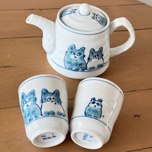 以下が含まれることがあります： 蓋付きのティーポットと2つの湯呑みがセットになった白い陶器の茶器セット。各アイテムには、大きな目をした猫の青い手描きイラストが施されています。ティーポットには横手ハンドルと注ぎ口が付いています。