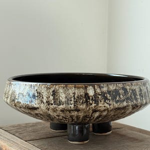 Può includere: Una grande ciotola in ceramica con un design ampio e poco profondo. La ciotola ha una superficie strutturata e screziata nei toni del marrone e crema, con un interno marrone scuro. È sostenuta da tre gambe corte e cilindriche. La ciotola è appoggiata su una superficie di legno.