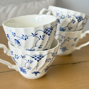 Può includere: Un set di tre tazze da tè in ceramica bianca con un delicato motivo floreale blu, viticci intrecciati e bordi smerlati. Ogni tazza presenta un manico curvo e un corpo scanalato, caratterizzato da una colorazione blu e bianca.