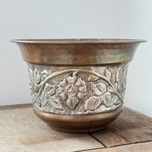Könnte beinhalten: Ein runder Pflanztopf aus Metall mit einem kupferfarbenen oberen Rand und Boden in gehämmertem Design. Das Mittelband besteht aus kontrastierendem silberfarbenem Metall mit einem detaillierten Reliefmuster aus Blumen und Ranken.