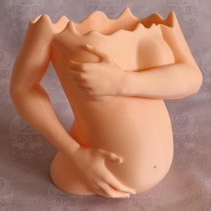 May include: A light peach-coloured ceramic planter shaped like a pregnant woman with her hands on her belly. The planter has a wavy top edge and is designed to hold plants or flowers.