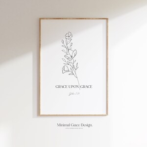 May include: A framed art print featuring a minimalist floral line drawing. The artwork is in black and white, with the words "GRACE UPON GRACE" and "John 1:16" printed below the floral design. The frame is a light brown color.