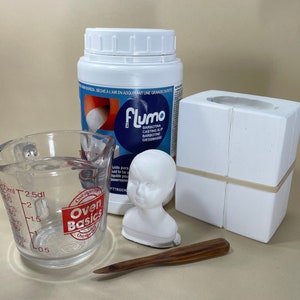 May include: A white plastic measuring cup with red markings, a white plastic container of casting slip, a white plastic mold for casting, a white plaster doll head, and a wooden tool.