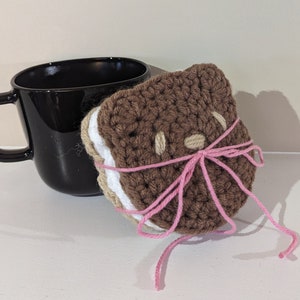May include: A handmade crochet coaster shaped like a sandwich, featuring brown, white, and tan yarn. The coaster is tied with pink yarn and sits next to a black mug. The coaster has tan circles for eyes and a tan oval for a nose.