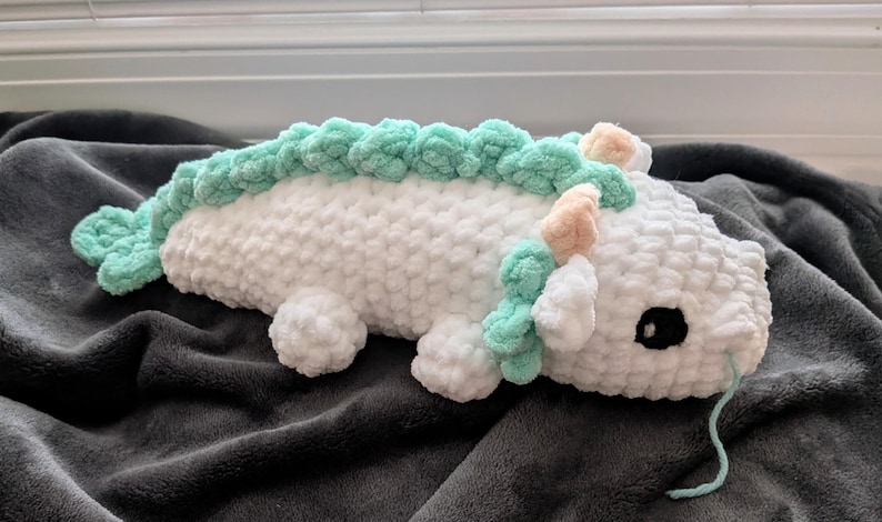May include: A handmade, white crochet dragon plushie with mint green accents along its back and tail. The dragon has small peach-colored horns and a black eye. The plushie is lying on a dark gray blanket.