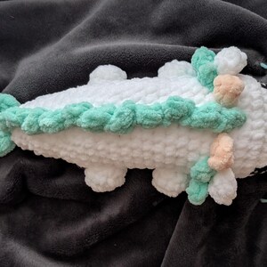 May include: A handmade white crochet creature with mint green and peach accents. The plush toy has a textured, fluffy appearance and is lying on a dark gray blanket. The creature has four small feet and a decorative border.