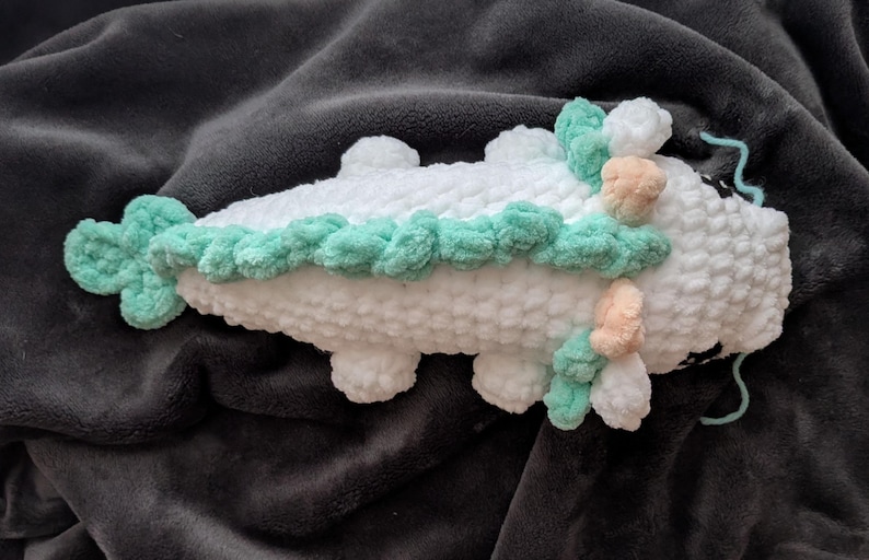 May include: A handmade white crochet creature with mint green and peach accents. The plush toy has a textured, fluffy appearance and is lying on a dark gray blanket. The creature has four small feet and a decorative border.