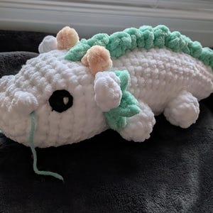 May include: A handmade white crochet dragon plushie with a green spine and accents. The dragon has black eyes, peach-colored horns, and light blue yarn whiskers. The plushie is lying on a dark gray blanket.