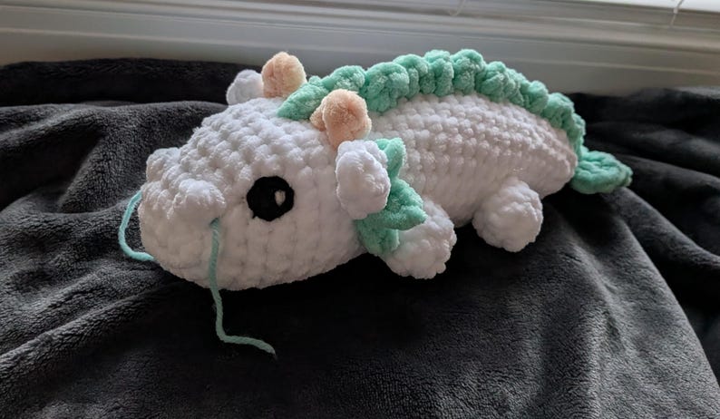 May include: A handmade white crochet dragon plushie with a green spine and accents. The dragon has black eyes, peach-colored horns, and light blue yarn whiskers. The plushie is lying on a dark gray blanket.