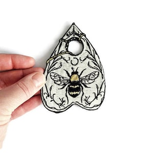 Puede incluir: Parche bordado con forma de planchette, con un diseño de abeja. La abeja tiene rayas negras y amarillas, con alas detalladas. El parche es de color crema con bordado negro y un borde decorativo.
