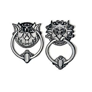 Può includere: Due battenti per porte in ferro ricamate in bianco e nero con un design dettagliato di un viso grottesco.