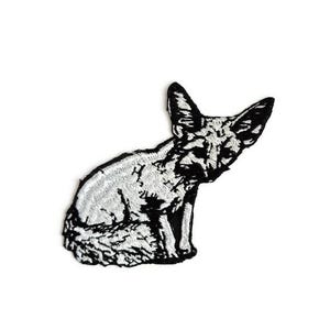 May include: Embroidered patch featuring a stylized fox in black and white. The fox is depicted in a seated position with its ears perked up. The patch is likely designed to be ironed or sewn onto fabric.