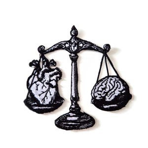 Pode incluir: Patch em preto e branco com uma balança. De um lado, um coração, do outro, um cérebro. O design sugere um tema de equilíbrio entre emoção e intelecto, com detalhes intrincados.
