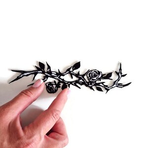 K&ouml;nnte beinhalten: Schwarzer floraler Zweig mit Rosen und Dornen. Das Design zeigt zwei Rosen und mehrere Bl&auml;tter. Das Bild befindet sich auf wei&szlig;em Hintergrund.