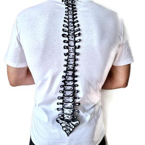 Puede incluir: Camiseta blanca con un dise&ntilde;o de columna vertebral en blanco y negro impreso en la espalda. La ilustraci&oacute;n de la columna vertebral es vertical. La camiseta tiene mangas cortas y cuello redondo.