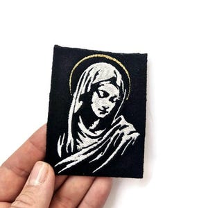 Könnte beinhalten: Weiß gesticktes Bild einer Frau mit Heiligenschein auf einem schwarzen Stoff-Patch.
