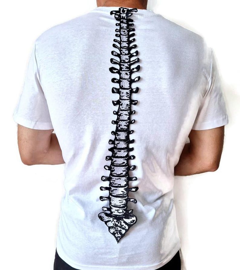 Puede incluir: Camiseta blanca con un dise&ntilde;o de columna vertebral en blanco y negro en la espalda. El dise&ntilde;o de la columna vertebral es detallado y recorre verticalmente el centro de la camiseta. La camiseta tiene mangas cortas y cuello redondo.