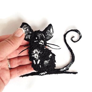 May include: Black embroidered cat patch with large ears and a curled tail. The cat is detailed with white accents and is set against a white background. This patch is suitable for clothing or accessories.